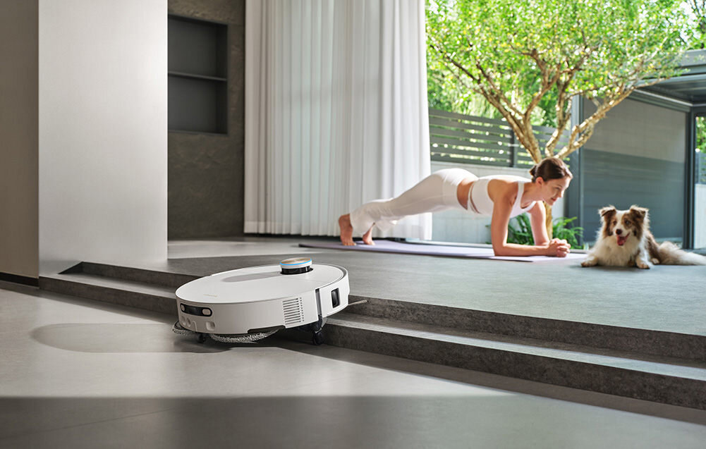 Biały robot sprzątający porusza się po podłodze w nowoczesnym wnętrzu. W tle osoba ćwiczy na macie, a obok niej siedzi pies.