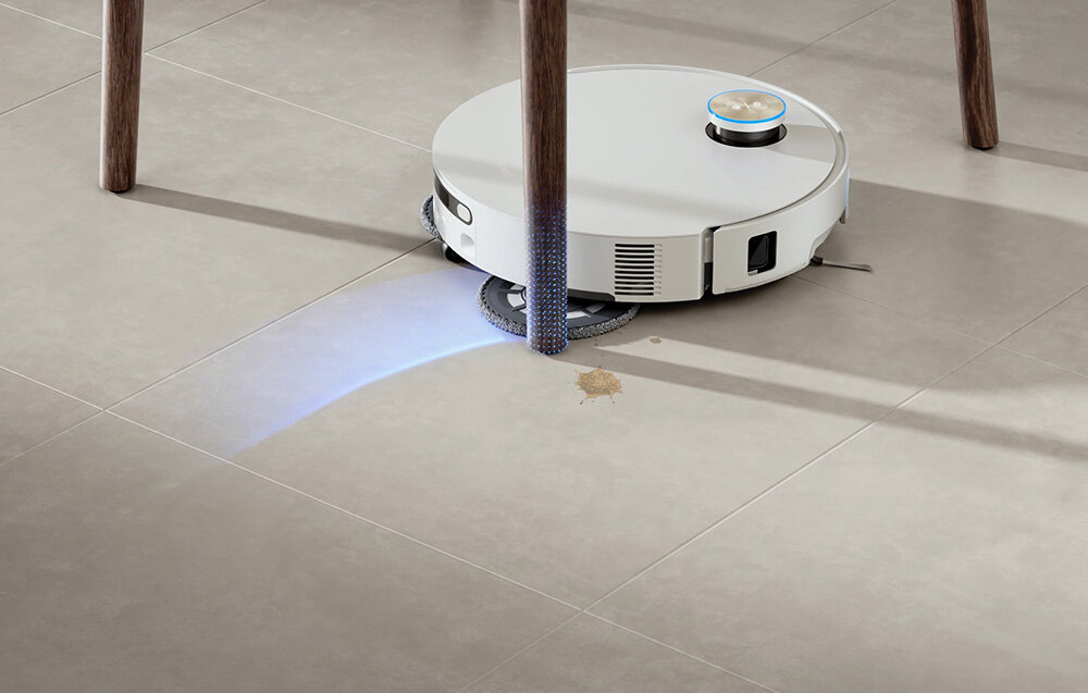 Biały robot sprzątający porusza się po jasnej podłodze, omijając nogę stołu. Na podłodze widoczna jest niewielka plama, a robot emituje niebieskie światło.