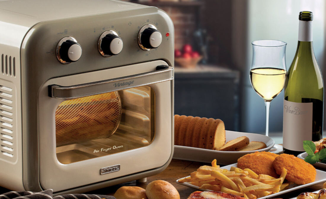 Beżowy mini piekarnik z trzema pokrętłami na górze i napisem \'Air Fryer Oven\' na drzwiczkach. Obok piekarnika znajdują się talerze z jedzeniem, kieliszek białego wina i butelka wina.