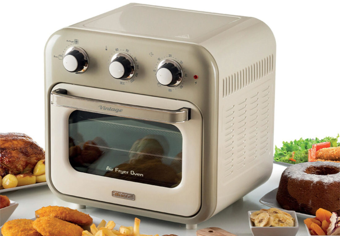 Beżowy mini piekarnik z trzema pokrętłami na górze i szklanymi drzwiczkami z napisem \'Air Fryer Oven\'. Wokół piekarnika znajdują się różne potrawy, w tym pieczone mięso, ciasto i warzywa.