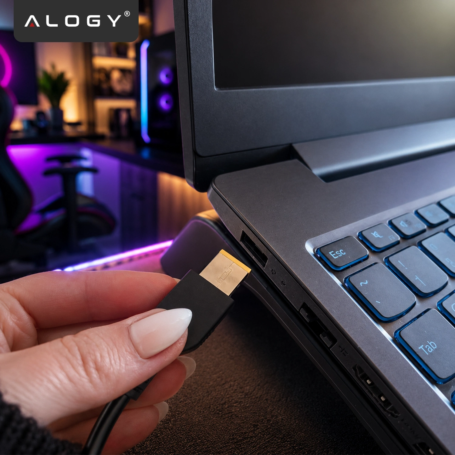 Osoba trzyma wtyczkę zasilacza, którą zbliża do gniazda laptopa. W tle widoczne jest biurko z komputerem i kolorowym oświetleniem LED, a w lewym górnym rogu znajduje się logo \'ALOGY\'.