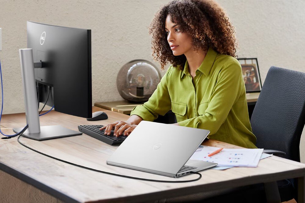 Osoba siedzi przy biurku, używając komputera stacjonarnego i laptopa z widocznym logo producenta. Na biurku znajdują się również dokumenty i długopisy.