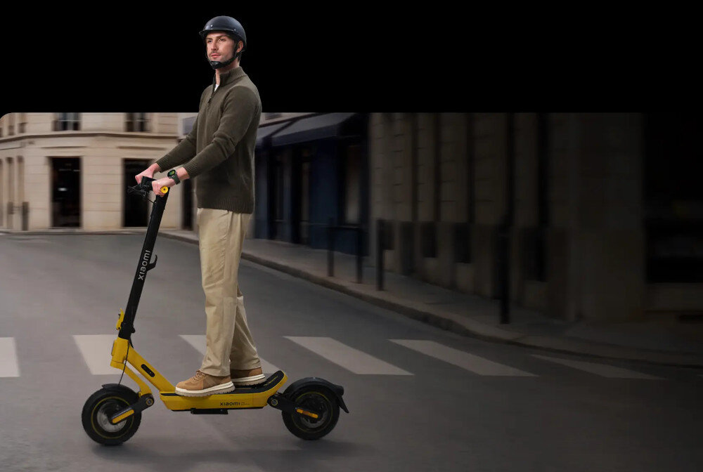 Osoba stoi na żółtej hulajnodze elektrycznej na ulicy miejskiej. Na hulajnodze widoczne jest logo \'Xiaomi\'.