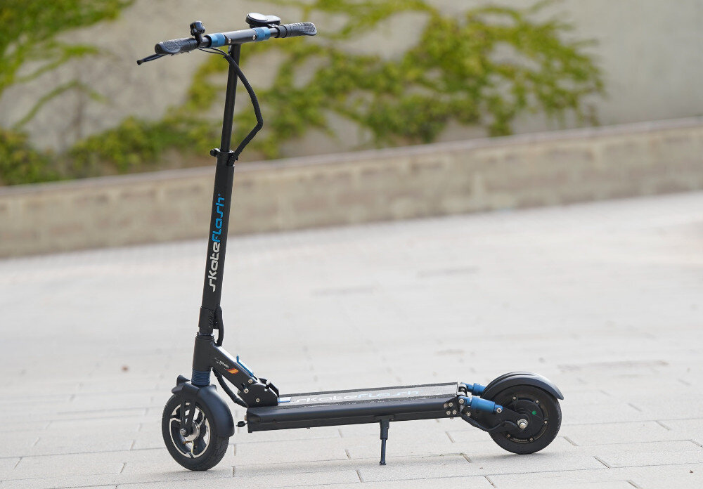 Czarna hulajnoga elektryczna z niebieskimi akcentami, stojąca na chodniku. Na kierownicy widoczny jest napis \'SkateFlash\'.