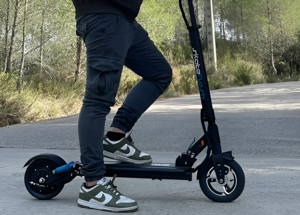 Osoba stoi na czarnej hulajnodze elektrycznej z widocznym napisem \'skateflash\' na kierownicy. W tle widać drogę i drzewa.