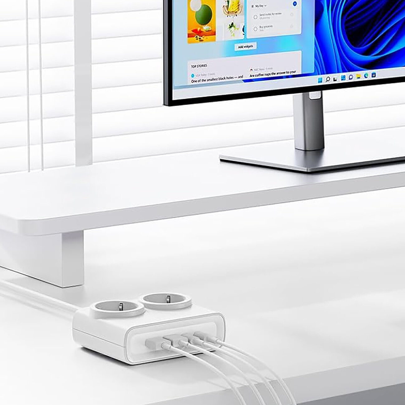 Na białym biurku znajduje się monitor oraz biała listwa zasilająca z dwoma gniazdami AC i czterema portami USB, do której podłączone są kable. W tle widać żaluzje na oknie.