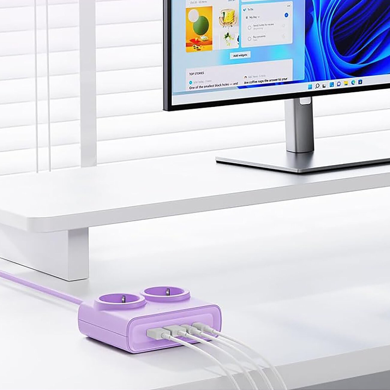 Fioletowa listwa zasilająca z dwoma gniazdami AC oraz czterema portami USB (dwa USB-A i dwa USB-C) leży na białym biurku. W tle widoczny jest monitor z wyświetlonym interfejsem systemu operacyjnego.
