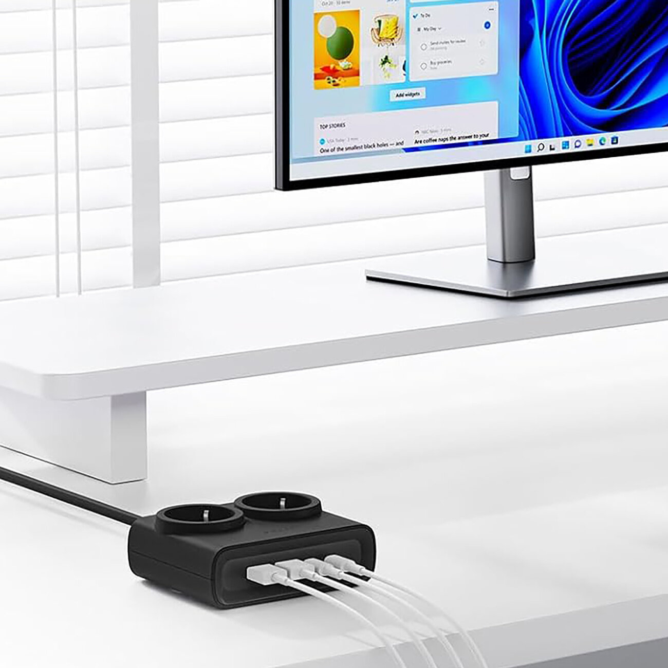 Na białym biurku znajduje się czarna listwa zasilająca z dwoma gniazdami AC i czterema portami USB, do której podłączone są białe kable. W tle widać monitor z wyświetlonym interfejsem systemu operacyjnego.