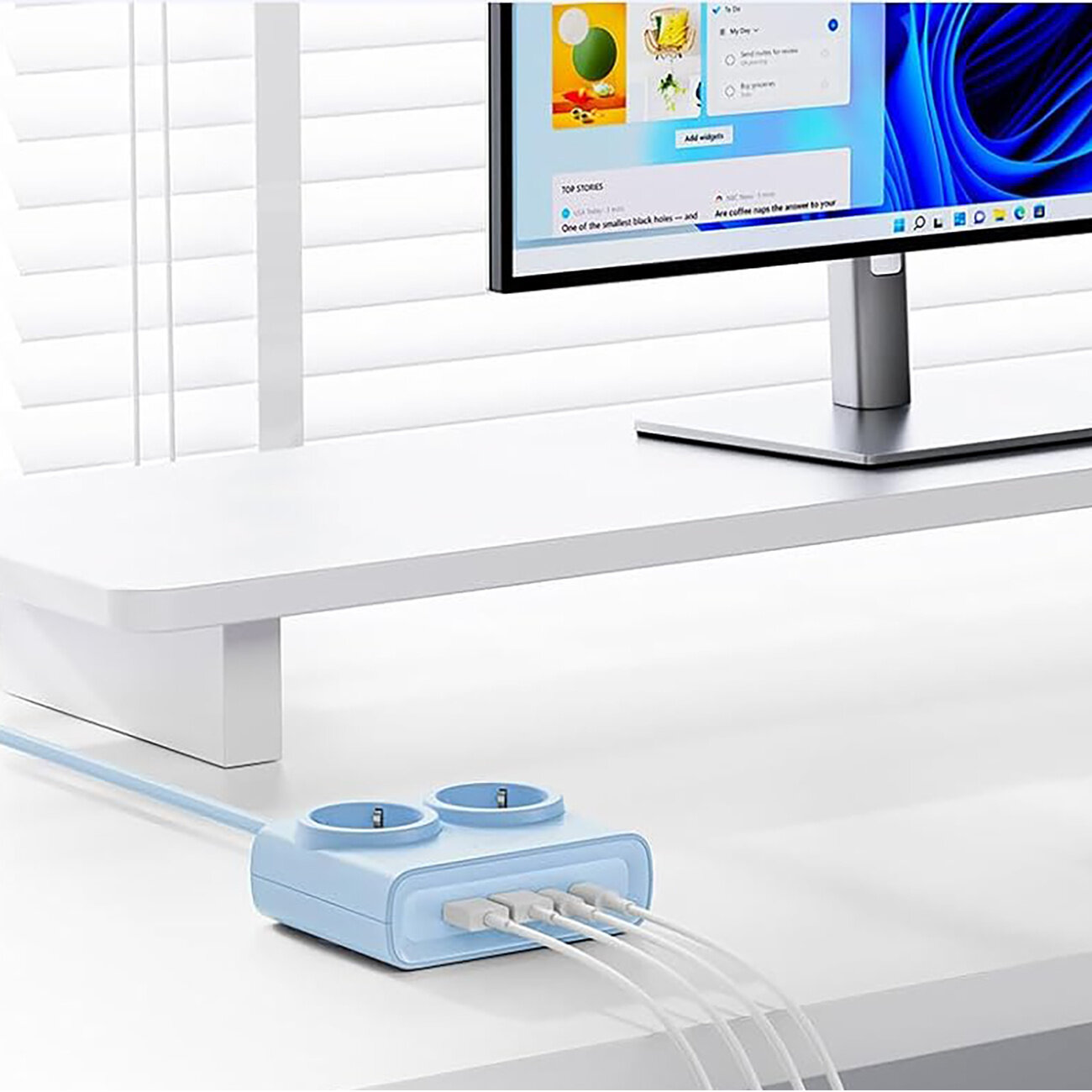 Na białym biurku znajduje się niebieska listwa zasilająca z dwoma gniazdami AC i czterema portami USB, do której podłączone są kable. Obok stoi monitor z wyświetlonym interfejsem systemu operacyjnego.