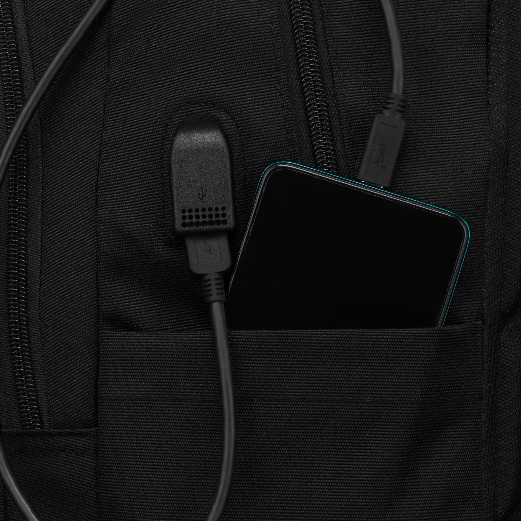 Czarny plecak z widocznym portem USB i kieszenią, w której znajduje się smartfon. Obok portu USB widać podłączony kabel.