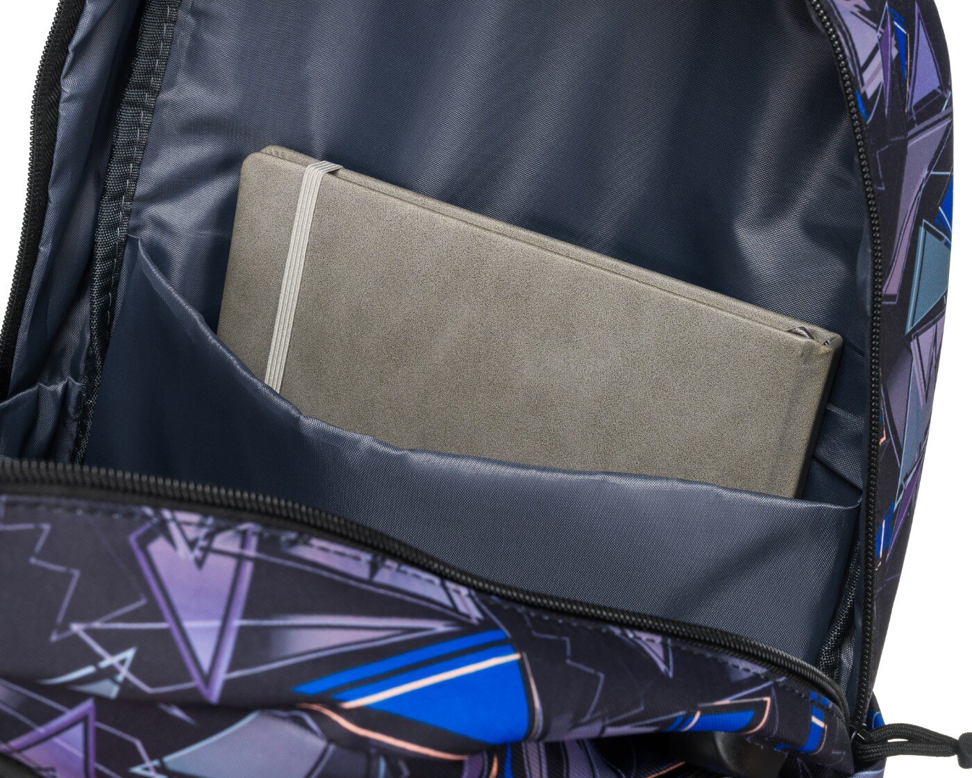 Wnętrze plecaka z geometrycznym wzorem, w którym znajduje się szara książka lub notes w kieszeni. Materiał plecaka jest ciemny z kolorowymi, geometrycznymi wzorami.