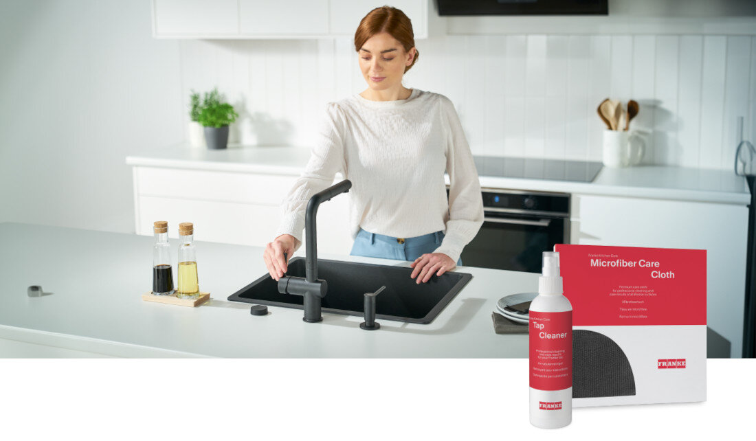 Kobieta stoi przy zlewie kuchennym z czarną matową baterią stojącą z wyciąganą wylewką. Obok widoczny jest zestaw do pielęgnacji z napisem \'Tap Cleaner\' i \'Microfiber Care Cloth\' z logo FRANKE.