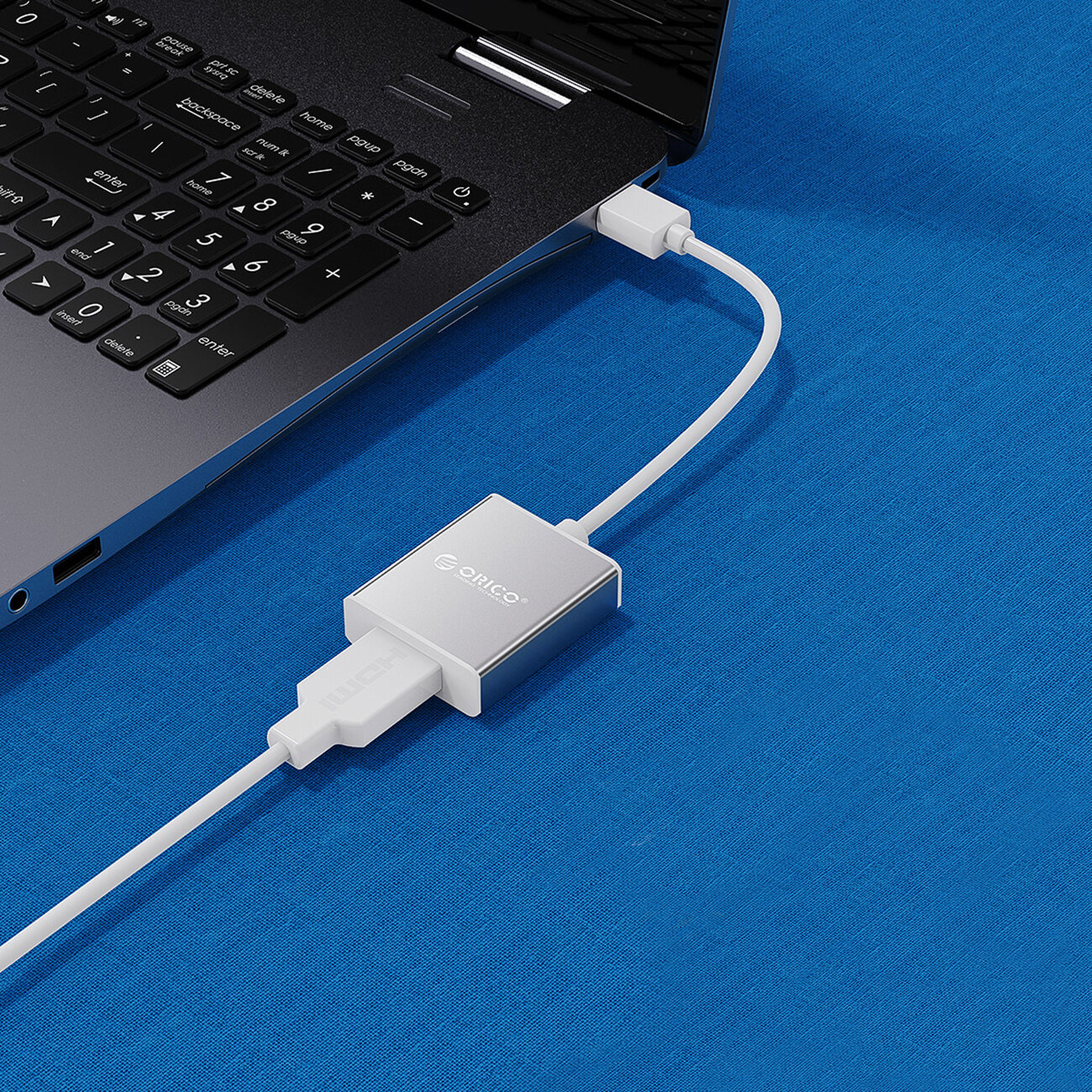 Na niebieskim tle znajduje się laptop z podłączonym srebrnym adapterem USB-A 3.0 do HDMI marki Orico. Adapter jest połączony białym kablem z laptopem.