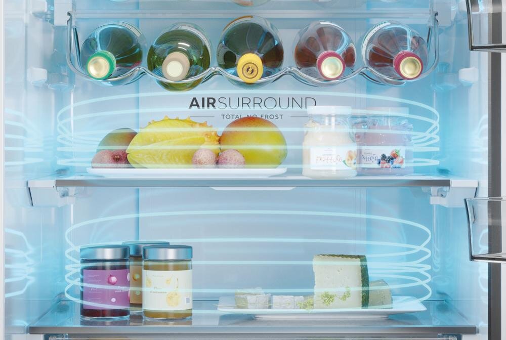 Wnętrze lodówki z półkami, na których znajdują się owoce, słoiki z przetworami i butelki wina. Na tylnej ściance widoczny jest napis \'AIR SURROUND TOTAL NO FROST\'.