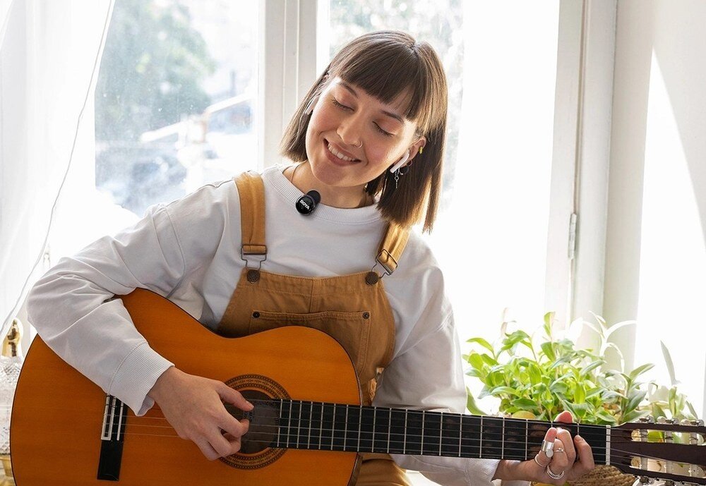 Osoba gra na gitarze akustycznej, mając przypięty mikrofon do ubrania. W tle widać okno i rośliny doniczkowe.