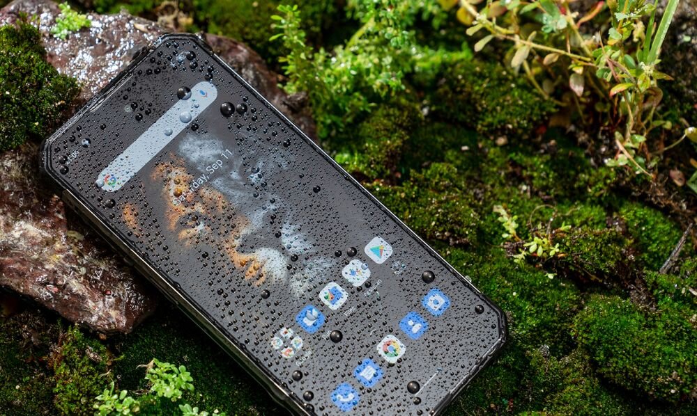 Smartfon leży na mokrej powierzchni pokrytej mchem, z widocznymi kroplami wody na ekranie. Na ekranie widać ikonki aplikacji oraz tekst \'1 Sep\'.