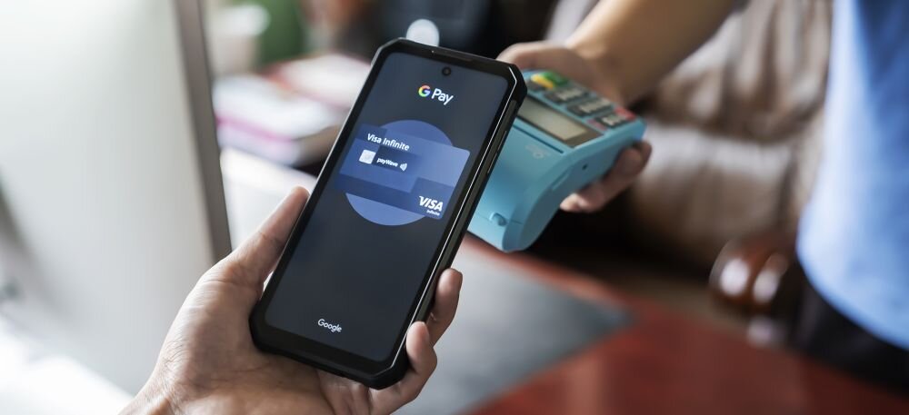 Osoba trzyma smartfon z aplikacją G Pay, na ekranie widoczna jest karta Visa Infinite. W tle znajduje się terminal płatniczy.