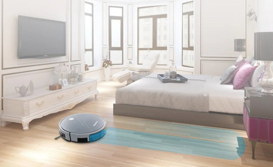 W elegancko urządzonym pokoju znajduje się robot sprzątający na podłodze, obok dużego łóżka z poduszkami. Na robocie widoczne jest logo producenta.