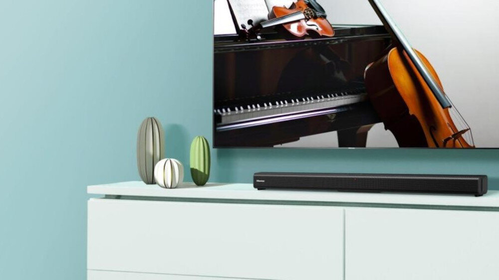Na białej szafce stoi czarny soundbar, a nad nim wisi telewizor z obrazem fortepianu i wiolonczeli. Obok soundbara znajdują się trzy dekoracyjne wazony w odcieniach zieleni i szarości.