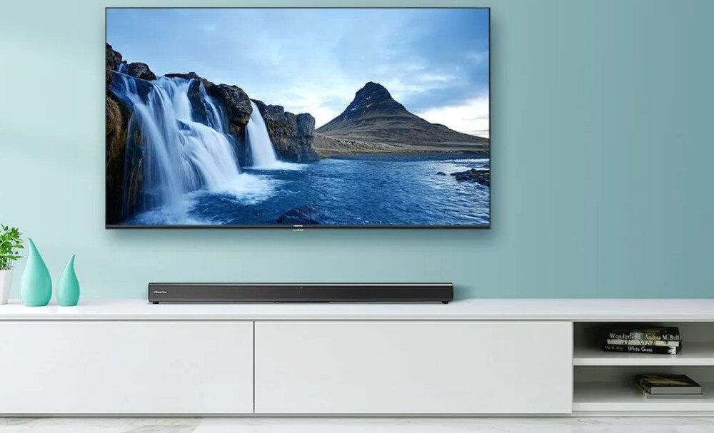 Na obrazie widać telewizor z wyświetlonym krajobrazem wodospadu i góry, umieszczony na jasnej szafce. Przed telewizorem znajduje się czarny soundbar, a obok szafki stoją trzy zielone wazony.