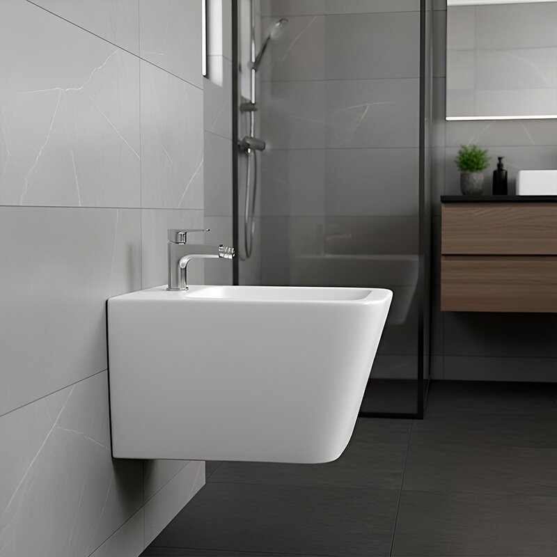 Na obrazie widoczny jest biały bidet zamontowany na ścianie w nowoczesnej łazience z szarymi płytkami. W tle znajduje się prysznic oraz drewniana szafka z umywalką.