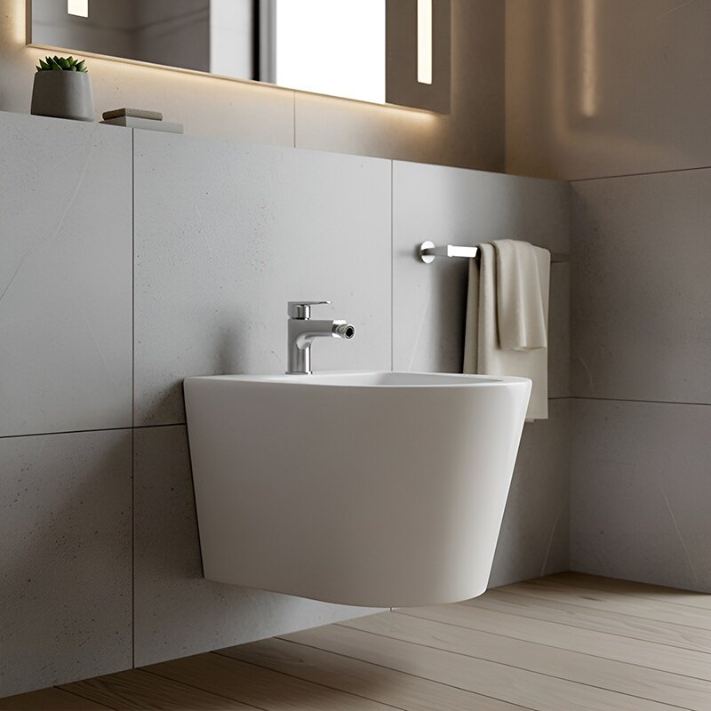 Na obrazie widoczny jest biały, podwieszany bidet łazienkowy z chromowaną baterią. W tle znajduje się ściana z jasnych płytek oraz ręcznik zawieszony na uchwycie.