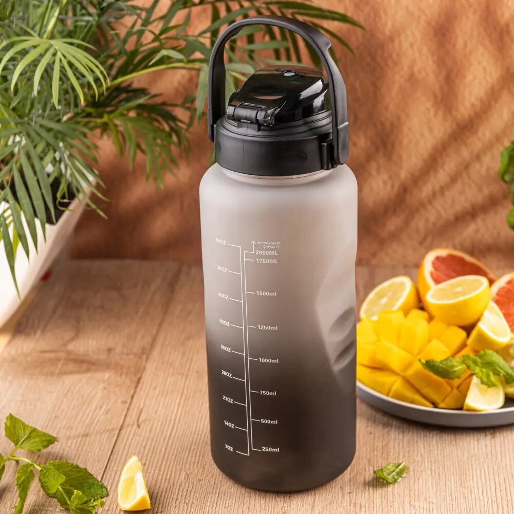 Na drewnianym stole stoi duża, szara butelka na wodę z czarną pokrywką i uchwytem, z podziałką do 2000 ml. Obok znajduje się talerz z pokrojonymi owocami, a w tle widać zieloną roślinę.