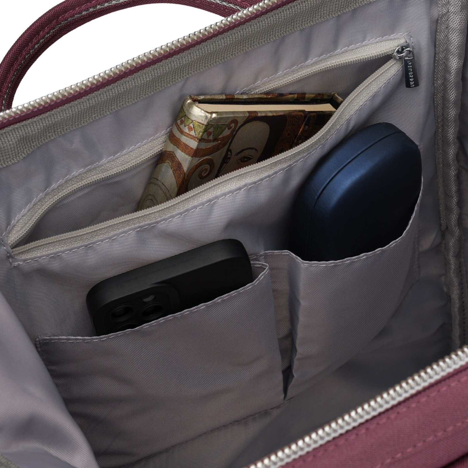 Wnętrze plecaka z kilkoma kieszeniami, w których znajdują się portfel z wzorem, czarne etui na telefon oraz niebieskie etui. Na zamku widoczny jest napis \'PETERSON\'.