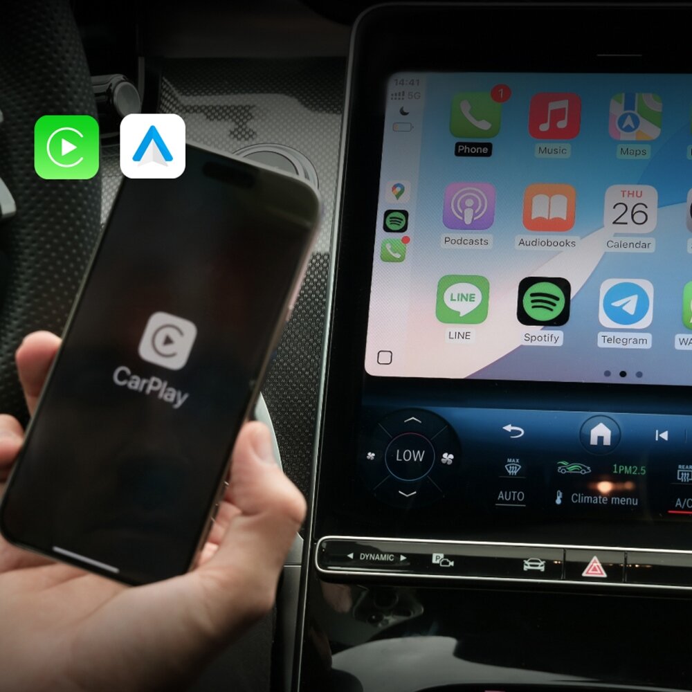 Osoba trzyma smartfon z włączoną aplikacją CarPlay, a obok znajduje się ekran samochodowy z interfejsem aplikacji, na którym widoczne są ikony takie jak Phone, Music, Maps, Podcasts, Audiobooks, Calendar, LINE, Spotify i Telegram. W górnym rogu ekranu widoczne są ikony Android Auto i CarPlay.