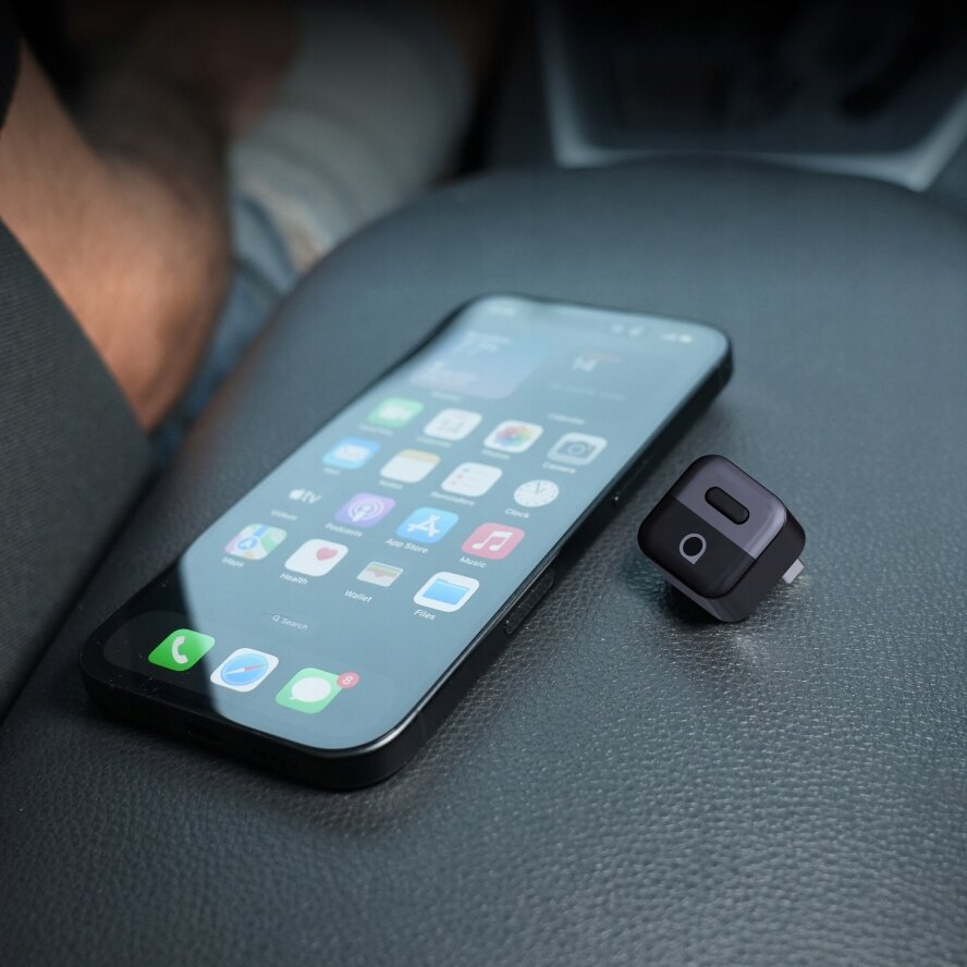 Na zdjęciu widać smartfon z wyświetlonymi ikonami aplikacji oraz małe, czarne urządzenie z logo \'a\' na górze, leżące na ciemnej powierzchni.