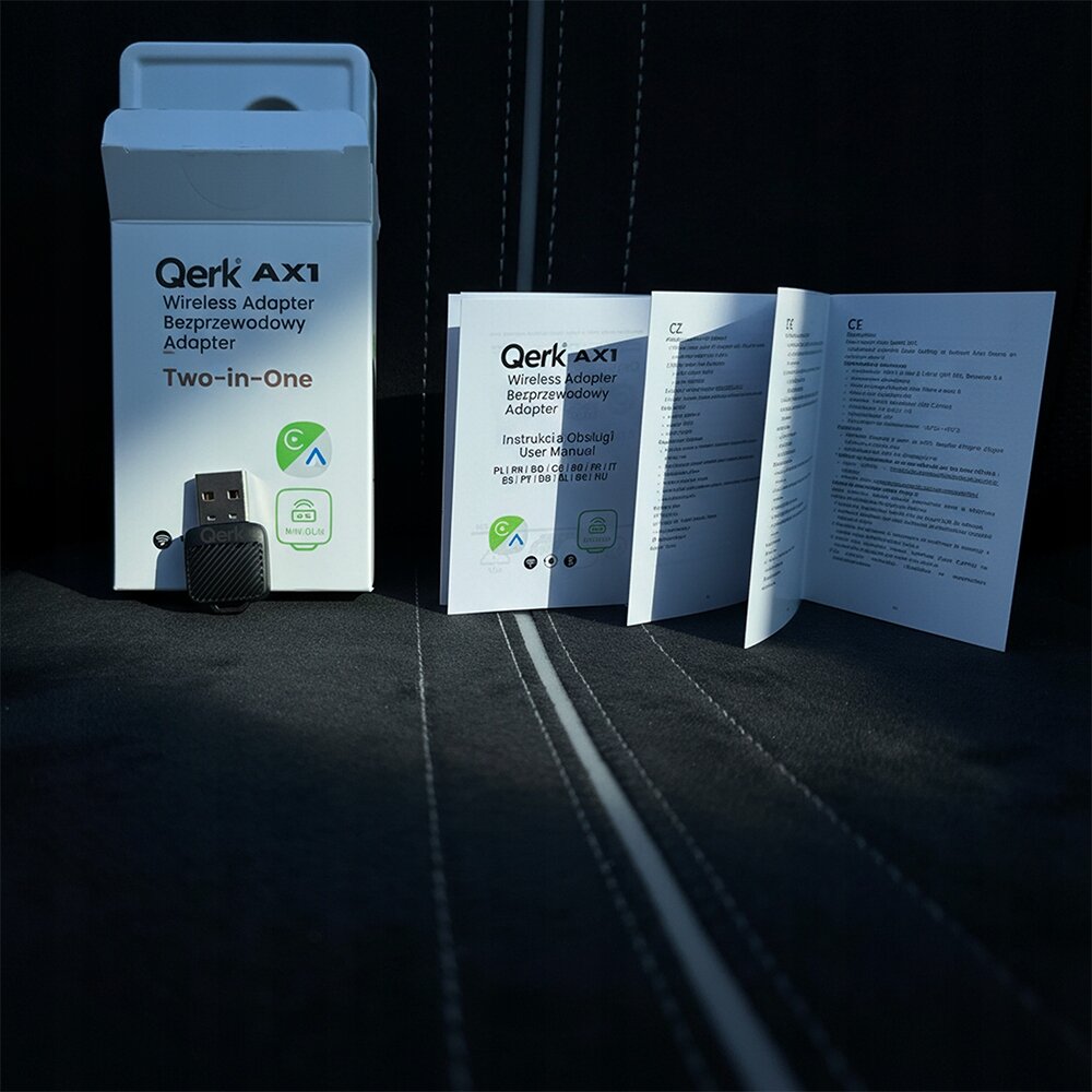 Na obrazie znajduje się opakowanie produktu Qerk AX1 Wireless Adapter Bezprzewodowy Adapter Two-in-One oraz instrukcja obsługi. Obok opakowania leży adapter USB z napisem \'Qerk\'.
