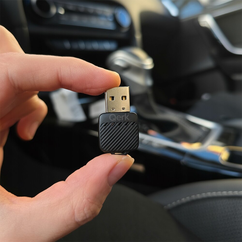Osoba trzyma w palcach mały adapter USB z napisem \'Qerk\' na czarnej obudowie. W tle widoczne jest wnętrze samochodu.