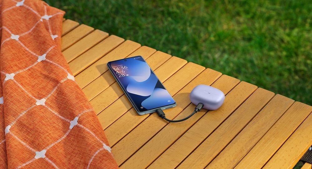 Na drewnianym stole leży smartfon z wyświetlaczem pokazującym godzinę 14:36, podłączony kablem do fioletowego etui na słuchawki. Obok znajduje się pomarańczowy materiał z białym wzorem, a w tle widoczna jest trawa.