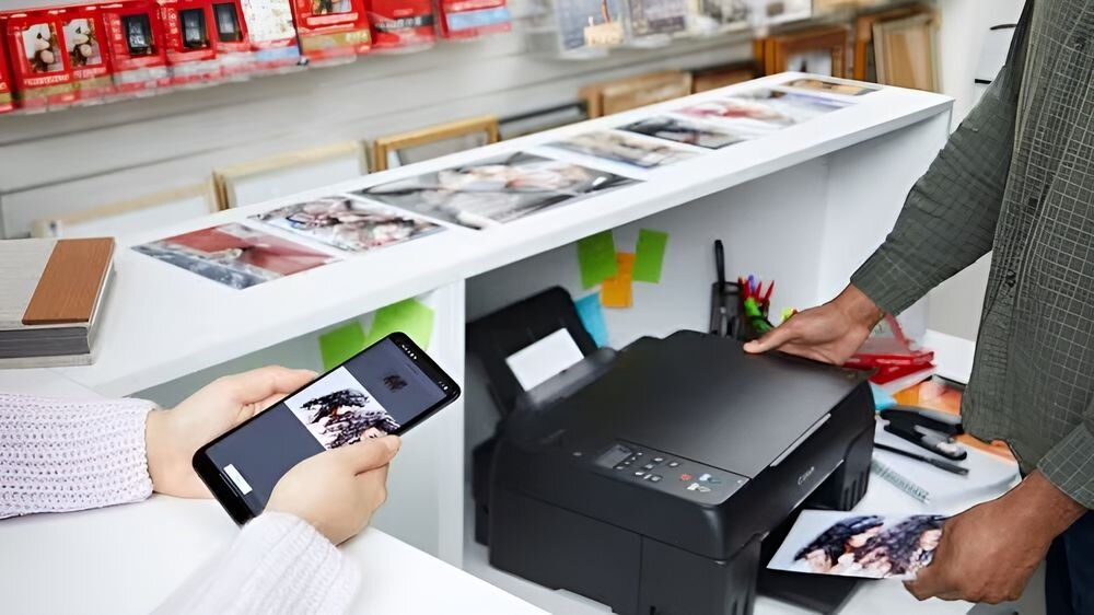 Na ladzie znajduje się drukarka, z której wychodzi kolorowy wydruk. Obok osoba trzyma smartfon z wyświetlonym zdjęciem.