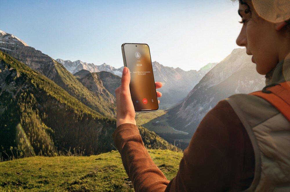Osoba trzyma smartfon z widocznym ekranem, na którym wyświetlany jest ekran połączenia przychodzącego. W tle rozciąga się malowniczy krajobraz górski z zielonymi dolinami i ośnieżonymi szczytami.