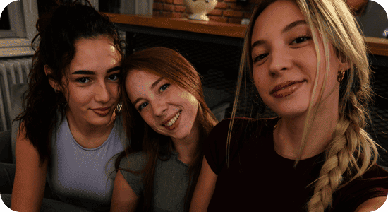 Trzy dziewczyny na kanapie robią selfie w słabym świetle; Nightography Galaxy A37 5G daje jasne, żywe detale. Trzy osoby uśmiechają się do aparatu w przytulnym wnętrzu z ceglaną ścianą w tle.