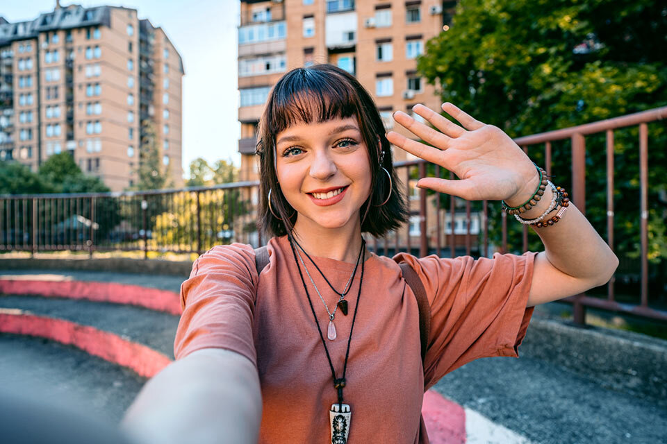 Osoba robiąca selfie na tle miejskiego krajobrazu z budynkami mieszkalnymi i zielenią. Osoba ma na sobie bransoletki i naszyjniki, a w tle widać barierki i schody.