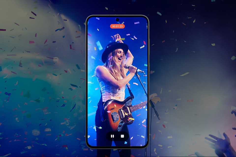 Na obrazie widać smartfon z wyświetlaczem pokazującym osobę grającą na gitarze na scenie, otoczoną konfetti. Na ekranie widoczny jest licznik czasu z napisem \'00:01:23\'.