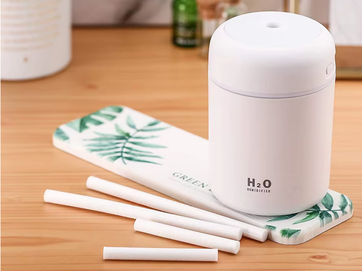 Na drewnianym blacie stoi biały nawilżacz powietrza z napisem \'H2O Humidifier\'. Obok leży kilka białych patyczków oraz podkładka z zielonym wzorem liści i napisem \'GREEN\'.