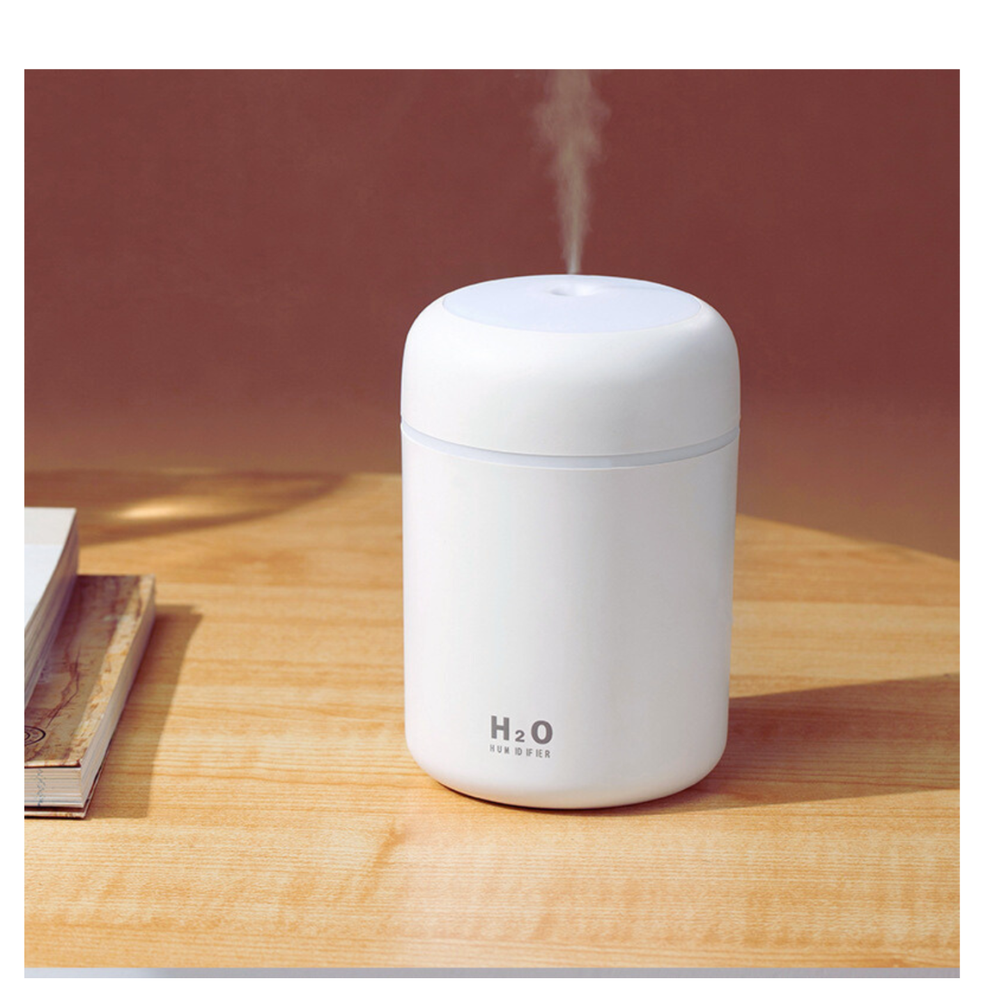 Na drewnianym blacie stoi biały nawilżacz powietrza, z którego unosi się delikatna mgiełka. Na urządzeniu widoczny jest napis \'H2O HUMIDIFIER\'.