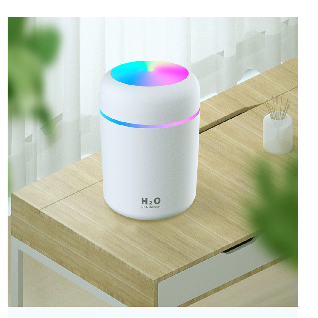Na drewnianym blacie stoi biały nawilżacz powietrza z kolorowym podświetleniem na górze. Na urządzeniu widoczny jest napis \'H2O Humidifier\'.