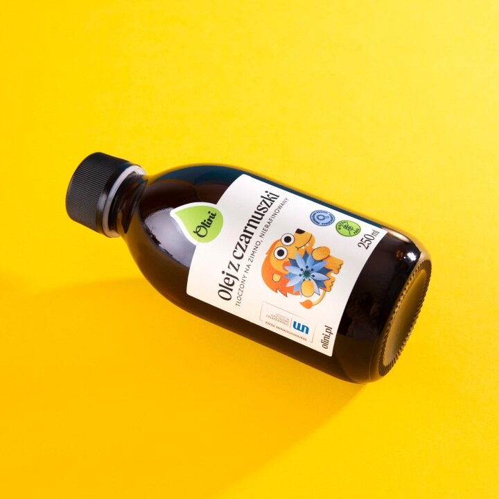 Butelka z ciemnego szkła z etykietą \'Olej z czarnuszki, tłoczony na zimno i nieprzetworzony, 250 ml\'. Na etykiecie znajduje się rysunek jeża trzymającego kwiat oraz logo \'Olini\'.
