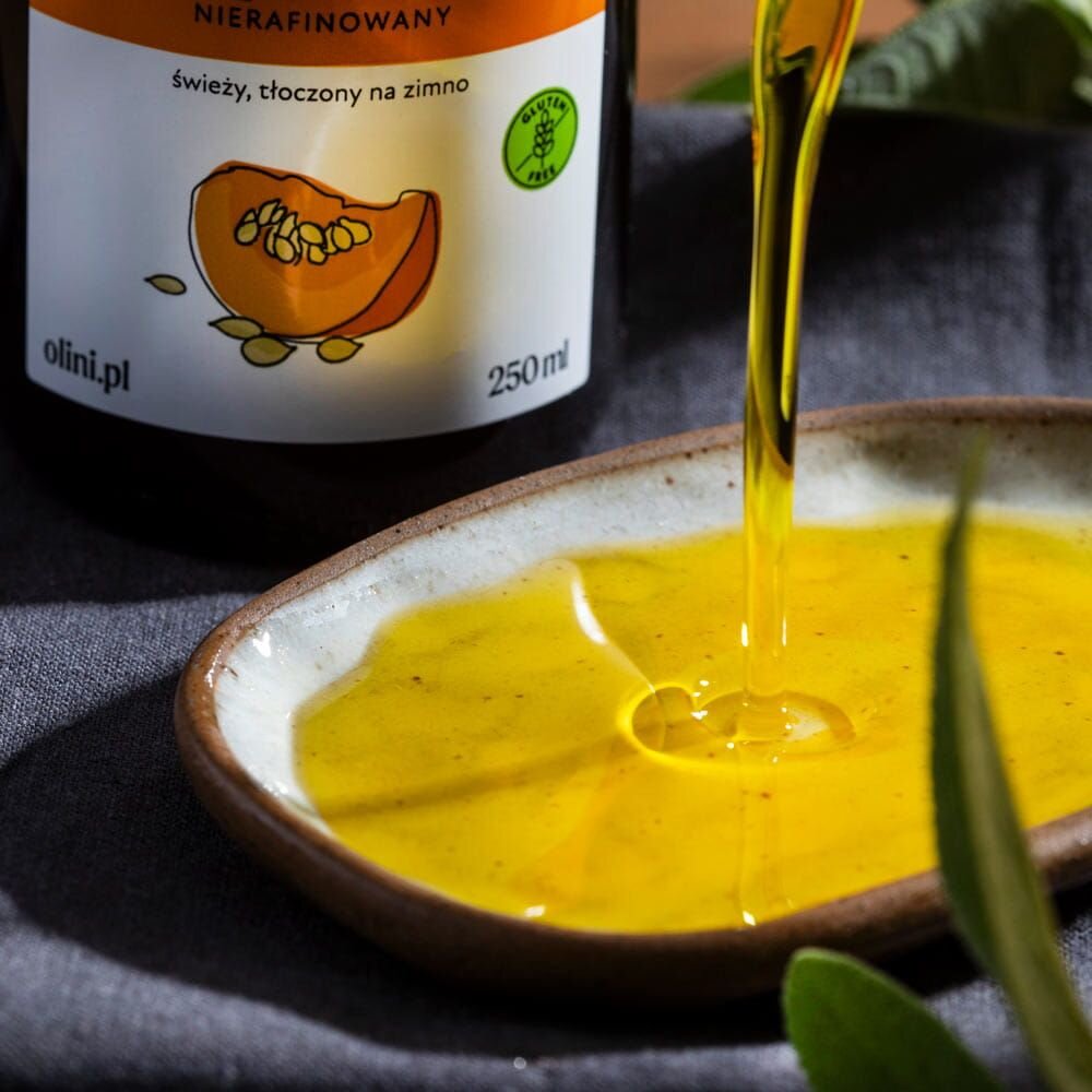 Na obrazie widoczna jest butelka z etykietą \'Olini Olej z Pestek Dyni 250 ml\', obok której olej wlewany jest na talerz. Etykieta zawiera ilustrację dyni oraz napisy \'świeży, tłoczony na zimno\' i \'gluten free\'.
