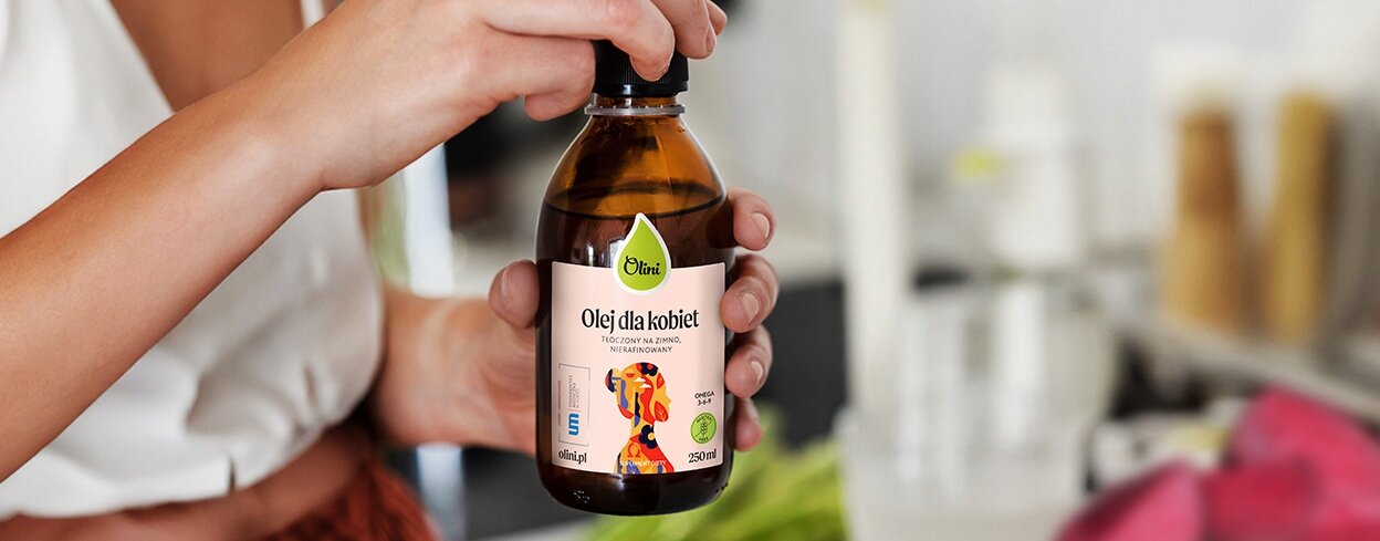 Osoba trzyma butelkę produktu \'Olini Olej dla Kobiet, na Hormony i Skórę, 250 ML\'. Etykieta jest kolorowa z grafiką i logo \'Olini\'.