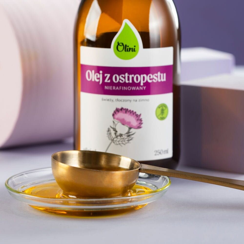 Na obrazie widoczna jest butelka z etykietą \'Olini Olej z ostropestu, nierafinowany, świeży, tłoczony na zimno, 250 ml\' oraz metalowa miarka na szklanym spodku z olejem.