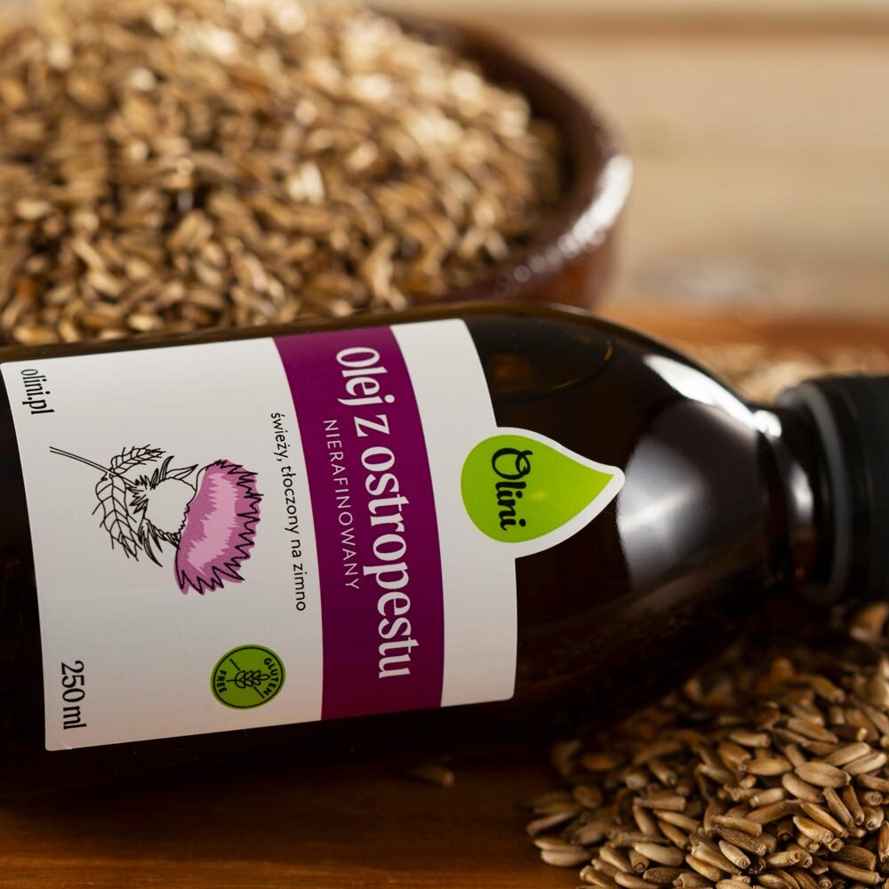 Butelka oleju z ostropestu marki Olini leży na tle nasion. Etykieta zawiera tekst: \'Olej z ostropestu, Nierafinowany, świeży, tłoczony na zimno, 250 ml\'.