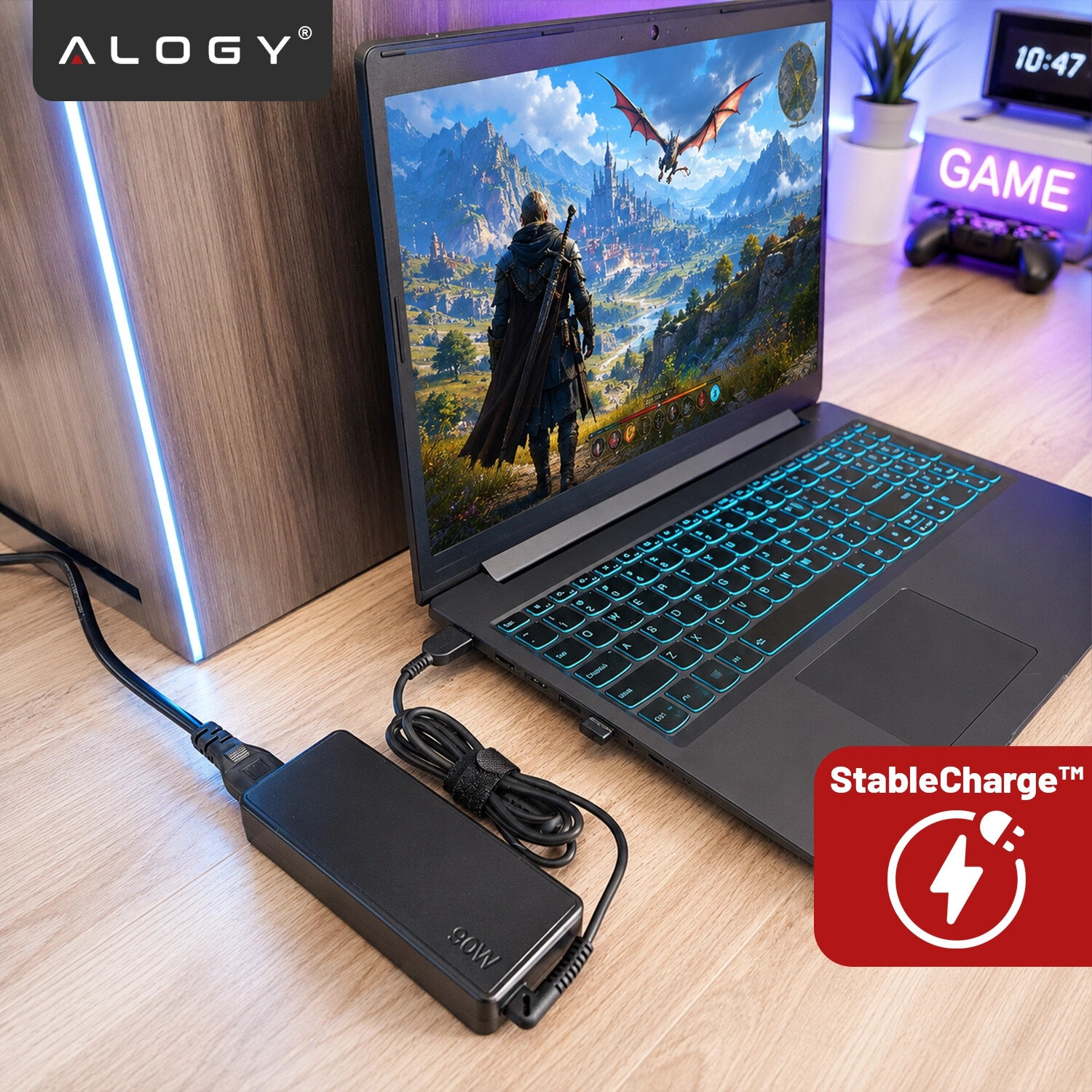 Na biurku znajduje się laptop z włączonym ekranem, na którym widoczna jest gra z postacią i smokiem. Obok laptopa leży czarny zasilacz z napisem \'90W\', a w rogu widoczne jest logo \'Alogy\' oraz ikona \'StableCharge™\'.