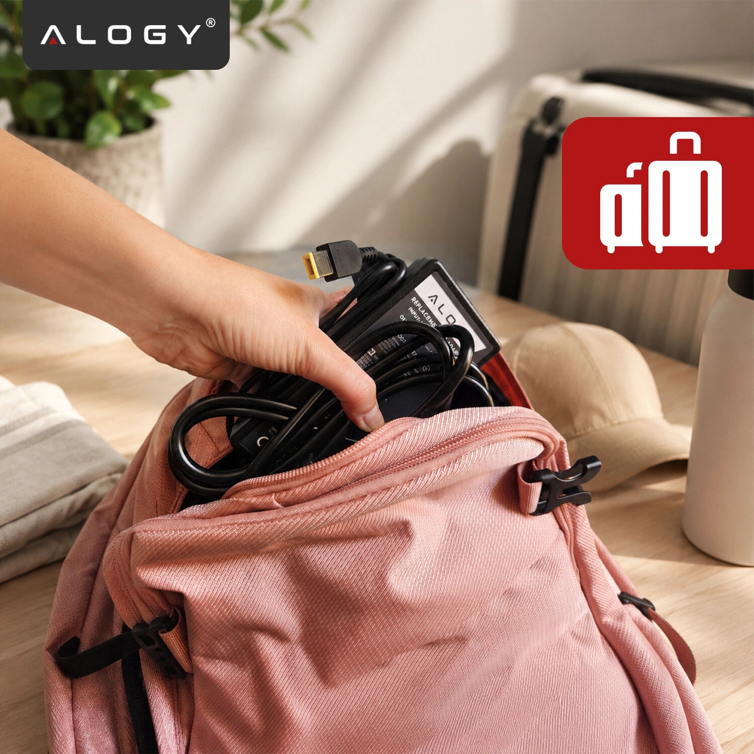 Osoba wkłada czarny zasilacz z logo \'ALOGY\' do różowego plecaka. W tle widoczne są kapelusz, butelka i roślina w doniczce.
