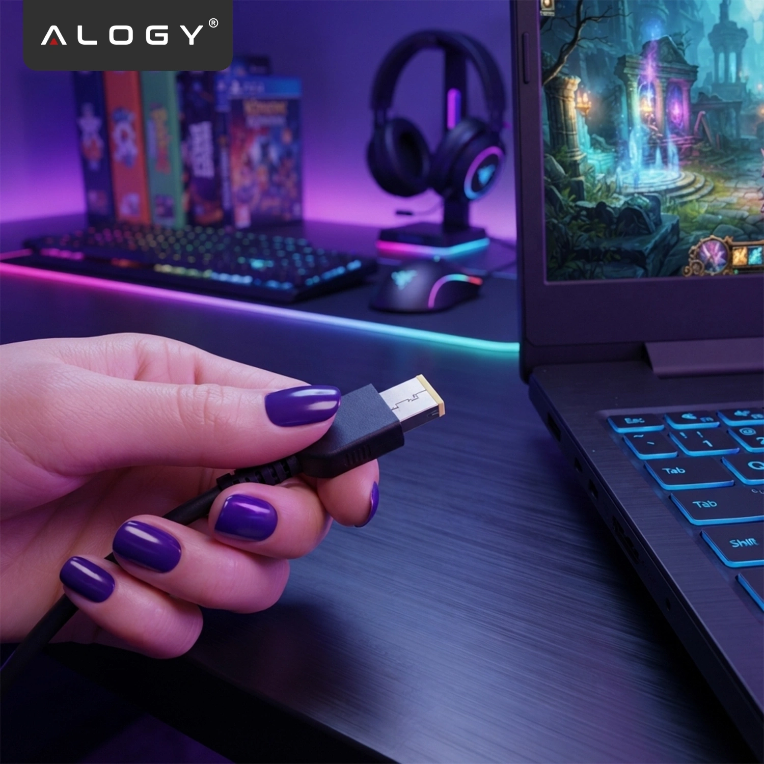 Osoba trzyma w dłoni czarny kabel z wtyczką, obok znajduje się laptop z podświetlaną klawiaturą. W tle widoczne są słuchawki, myszka i klawiatura z kolorowym podświetleniem oraz logo \'ALOGY\'.