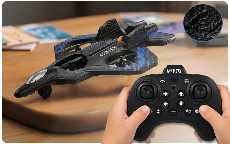 Na drewnianym stole znajduje się czarny dron w kształcie samolotu z napisem \'W26-X8\' i niebieskimi elementami. Przed dronem widoczny jest czarny kontroler z przyciskami, trzymany w dłoniach.